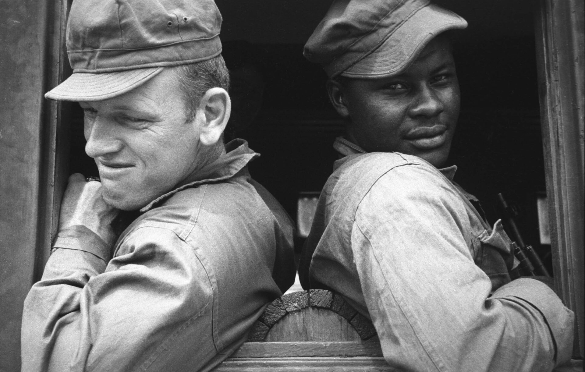 Two soldiers, one Black, one white back to back looking out train window headed to the front in the Korean War.