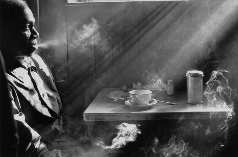 An man sits in a diner smoking as the smoke curls and wafts in the sunlight coming through a window,